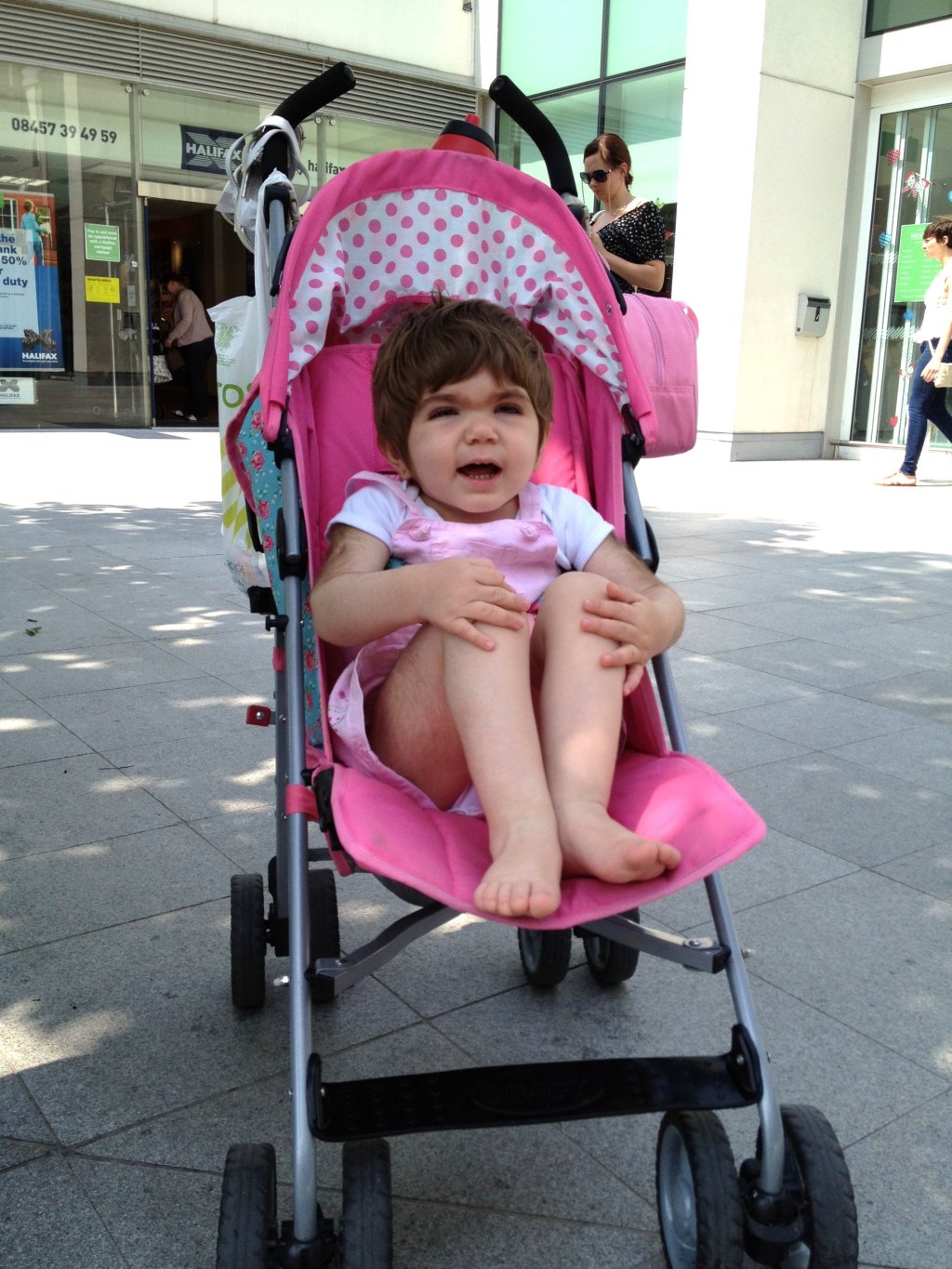 A young girl with MPS I Hurler sits in a pink pram with her knees tucked into her chest and an open smile on her face.