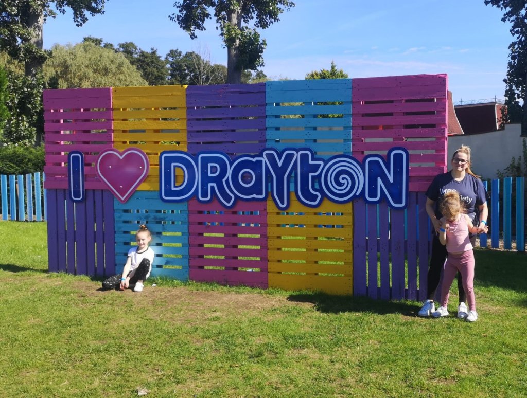 A family stand in front of the I heart Drayton Manor sign which is pink, blue, yellow and purple.