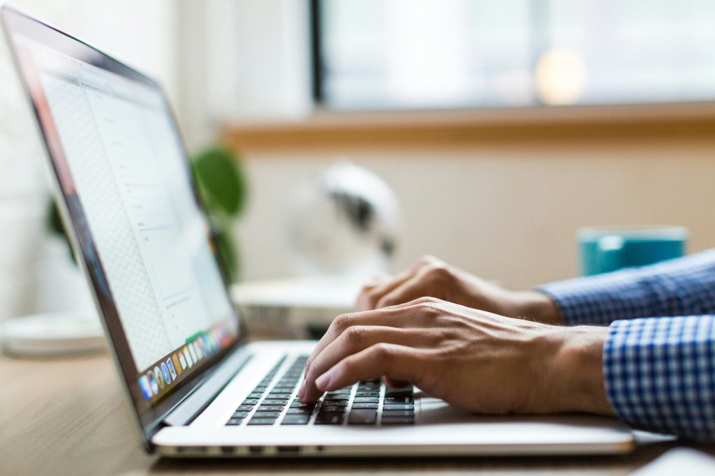 Hands of a man wearing a blue and white checked shirt on a MacBook laptop. Photo by Burst on Unsplash.